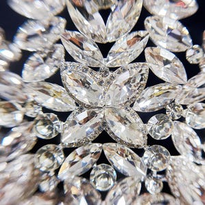 May include: Close-up of a sparkling, ornate crystal embellishment. The design features a central flower-like arrangement surrounded by numerous clear, faceted crystals of varying shapes and sizes, creating a dazzling effect. The crystals are set against a dark background.
