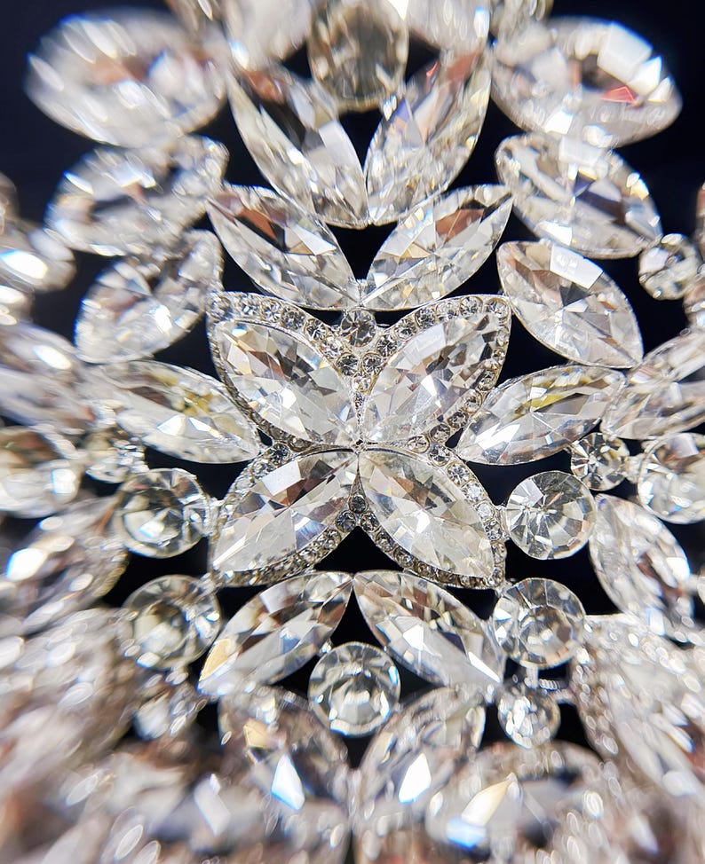May include: Close-up of a sparkling, ornate crystal embellishment. The design features a central flower-like arrangement surrounded by numerous clear, faceted crystals of varying shapes and sizes, creating a dazzling effect. The crystals are set against a dark background.