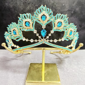 May include: A gold-colored tiara with turquoise and clear gemstones. The tiara features a peacock feather design with large turquoise teardrop-shaped stones and smaller clear stones. The tiara is displayed on a gold stand.