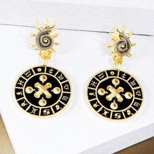 Könnte beinhalten: Goldfarbene Ohrringe mit schwarzen runden Anhängern mit Tierkreiszeichen und einem goldenen Kreuz. Die Ohrringe haben oben ein Spiraldesign.