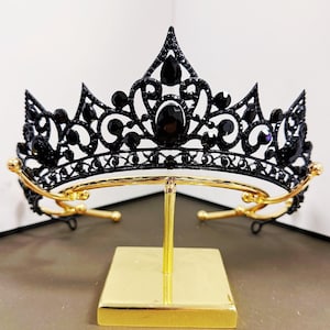 May include: A black crown with ornate detailing and black gemstones. The crown is displayed on a gold-colored stand with a square base. The crown's design features pointed arches and curved accents.