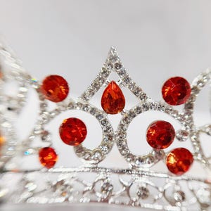 May include: A silver-toned metal tiara featuring a series of circular and teardrop-shaped orange crystals. The frame is intricately detailed with small clear rhinestones set throughout the metal structure, creating a sparkling, ornate headpiece.