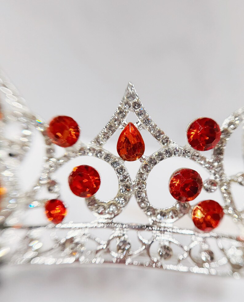 May include: A silver-toned metal tiara featuring a series of circular and teardrop-shaped orange crystals. The frame is intricately detailed with small clear rhinestones set throughout the metal structure, creating a sparkling, ornate headpiece.