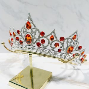 May include: A silver-toned tiara featuring intricate metal scrollwork and numerous clear rhinestones. The crown is embellished with multiple orange, oval, and round-cut faceted stones arranged in a symmetrical pattern across the front.