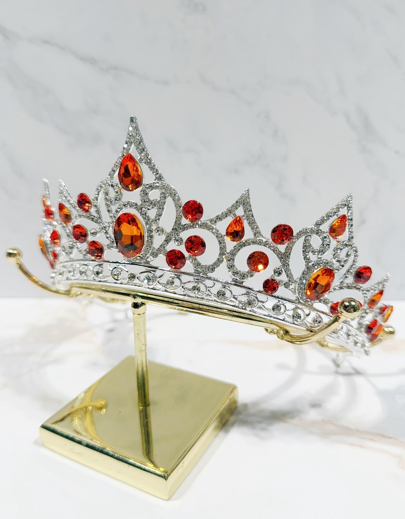 May include: A silver-toned tiara featuring intricate metal scrollwork and numerous clear rhinestones. The crown is embellished with multiple orange, oval, and round-cut faceted stones arranged in a symmetrical pattern across the front.