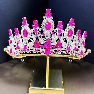 May include: A silver crown with pink rhinestones and a gold stand. The crown features a floral design and is decorated with teardrop-shaped stones.