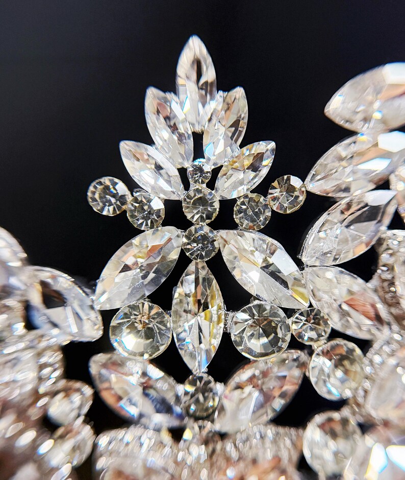May include: Close-up of a sparkling tiara with clear, faceted crystals. The tiara features a floral design with various shapes of crystals, including marquise and round cuts. The crystals are set in a silver-toned metal, creating a glamorous look.