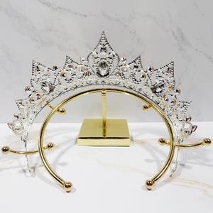 May include: A silver-toned metal tiara featuring intricate filigree patterns and clear embedded crystals, displayed on a gold-colored metal stand with a square base and a curved support frame with rounded ends for holding the headpiece securely.