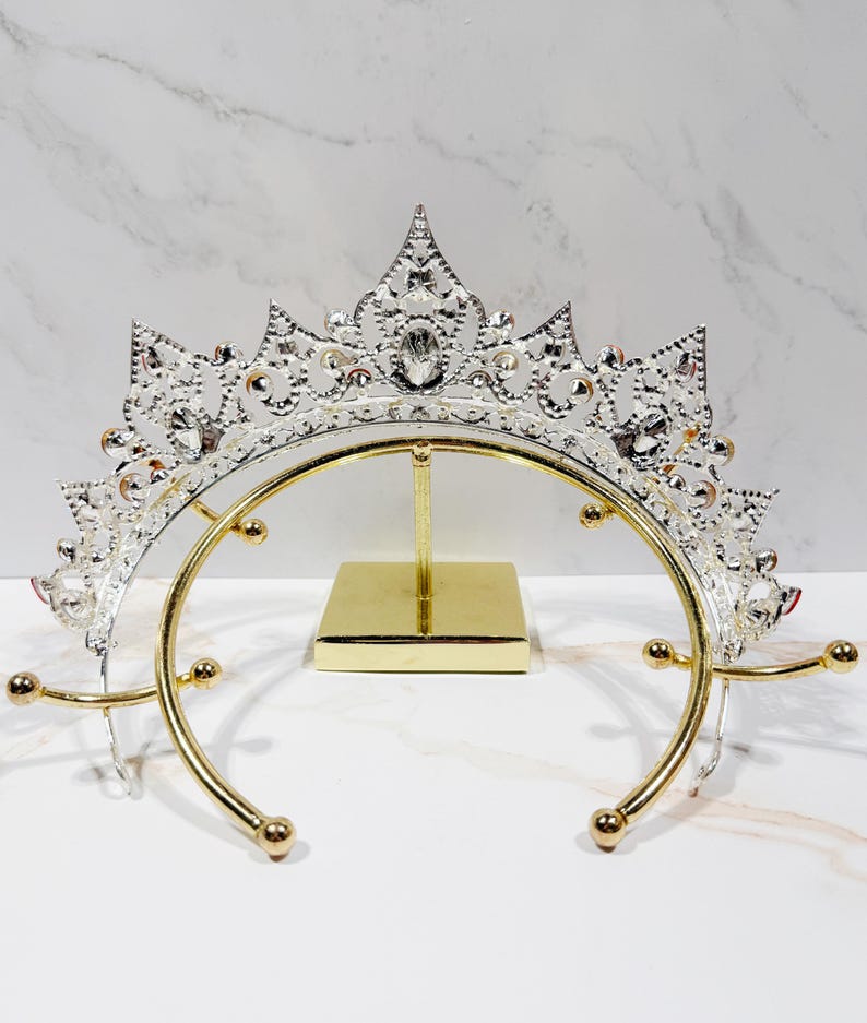 May include: A silver-toned metal tiara featuring intricate filigree patterns and clear embedded crystals, displayed on a gold-colored metal stand with a square base and a curved support frame with rounded ends for holding the headpiece securely.