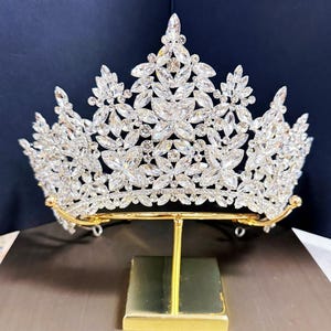 May include: A silver-toned crown with a floral design, adorned with numerous clear crystals. The crown is displayed on a gold-colored stand, set against a dark blue backdrop. The tiara's intricate details and sparkling stones create a luxurious appearance.