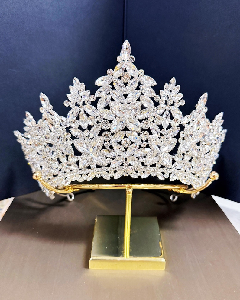 May include: A silver-toned crown with a floral design, adorned with numerous clear crystals. The crown is displayed on a gold-colored stand, set against a dark blue backdrop. The tiara's intricate details and sparkling stones create a luxurious appearance.