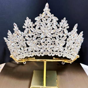 Könnte beinhalten: Eine funkelnde silberne Tiara mit klaren Kristallakzenten. Die Krone wird auf einem goldfarbenen Ständer präsentiert. Die Tiara zeigt ein florales Design mit zahlreichen Kristallverzierungen, perfekt für besondere Anlässe.