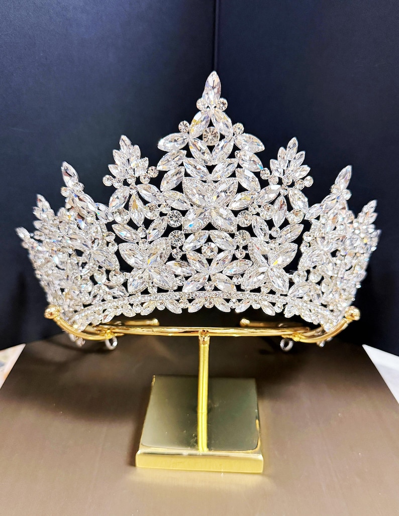 May include: A sparkling silver tiara with clear crystal accents. The crown is displayed on a gold-colored stand. The tiara features a floral design with multiple crystal embellishments, perfect for special occasions.