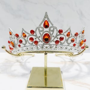 May include: A silver-toned tiara featuring a symmetrical, ornate metal framework encrusted with small clear crystals and accented by multiple vibrant orange, teardrop-shaped and round faceted stones arranged in a decorative, elevated pattern.