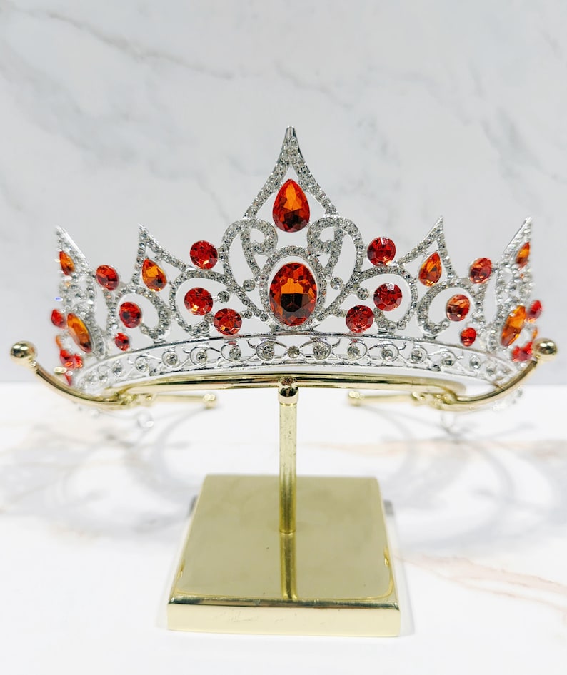 May include: A silver-toned tiara featuring a symmetrical, ornate metal framework encrusted with small clear crystals and accented by multiple vibrant orange, teardrop-shaped and round faceted stones arranged in a decorative, elevated pattern.