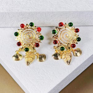 May include: A pair of gold-coloured earrings. Each earring has a sunburst design with a spiral centre, decorated with red and green beads. Each earring dangles a shell and a pinecone charm.