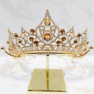 Peut inclure: Un diadème en métal doré présentant un motif complexe de volutes orné de nombreux strass blancs transparents et de plusieurs cristaux ambrés plus grands, de forme ovale et en goutte, monté sur un présentoir doré.