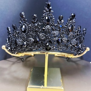May include: A black crystal crown with a gold-colored base. The crown is adorned with numerous black crystals of varying sizes and shapes, creating a sparkling effect. The crown is displayed on a gold-colored stand.