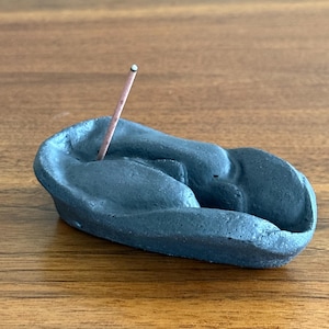 May include: A dark gray ceramic incense holder with a curved, organic shape. A lit incense stick is placed in the holder. The holder is on a wooden surface.