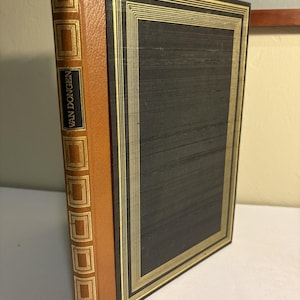 Puede incluir: Un libro de tapa dura con una cubierta gris oscuro y ribete dorado. El lomo es de color marrón claro con detalles dorados y el nombre "VAN DONGEN" impreso verticalmente. El libro está de pie sobre una superficie blanca.