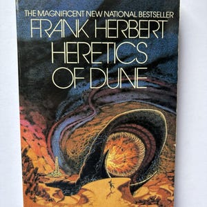 May include: A paperback book titled "Heretics of Dune" by Frank Herbert. The cover features a dark blue and orange illustration of a desert landscape with a large worm-like creature and the book's title in white.