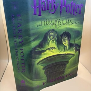 Puede incluir: Un libro de tapa dura titulado "Harry Potter y el Misterio del Príncipe". La portada presenta una combinación de colores verde y morado con ilustraciones de Dumbledore y Harry Potter. Se puede ver el nombre de la autora, J.K. Rowling.