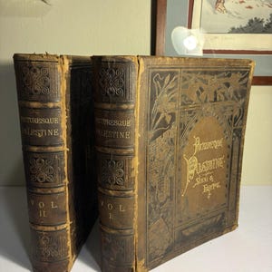 May include: Two antique leather-bound books, "Picturesque Palestine, Sinai & Egypt," with gold lettering and decorative embossed designs. Volume II and Volume I are visible. The books are brown with signs of wear.