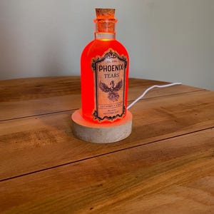 May include: A glowing orange bottle with a cork stopper, labeled "Phoenix Tears" with a phoenix illustration. The bottle sits on a wooden base and is lit from within. A white cord extends from the base.