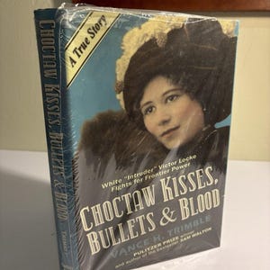 Può includere: Un libro intitolato "Choctaw Kisses, Bullets & Blood" di Vance H. Trimble. La copertina presenta una donna con un cappello e il testo "A True Story". Il libro è avvolto in plastica trasparente.