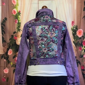 May include: Purple denim jacket with a sheer, sequined floral design on the back. The jacket has a cropped fit and a button closure.