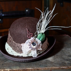 May include: A brown felt fedora hat with a lace trim and a steampunk-inspired gear and feather embellishment.
