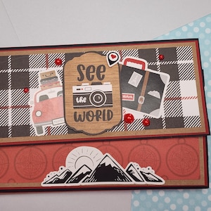 May include: A travel-themed card with a red and black plaid pattern. The card features illustrations of a red car with luggage, a camera, and a suitcase. The words "See the World" are printed on a wooden sign. Red embellishments add detail.