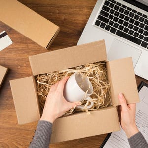 May include: A white ceramic mug being carefully removed from a cardboard box filled with packing material. The box is open, revealing the mug nestled inside. A laptop and other shipping supplies are visible on a wooden surface.