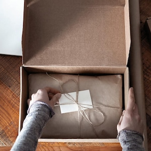 May include: A brown cardboard box with the lid open, revealing a package wrapped in brown paper and tied with twine. A small white tag is attached to the twine. The box is on a wooden surface.