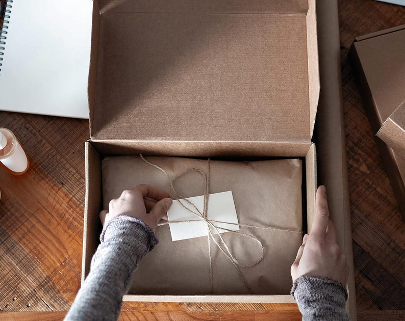 May include: A brown cardboard box with the lid open, revealing a package wrapped in brown paper and tied with twine. A small white tag is attached to the twine. The box is on a wooden surface.