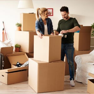 May include: A couple packing cardboard moving boxes in a room. The boxes are stacked, some open, and some are being taped shut. The room has a neutral color palette with a white ceiling and walls. Plants in terracotta pots are visible.