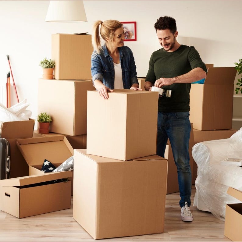 May include: A couple packing cardboard moving boxes in a room. The boxes are stacked, some open, and some are being taped shut. The room has a neutral color palette with a white ceiling and walls. Plants in terracotta pots are visible.