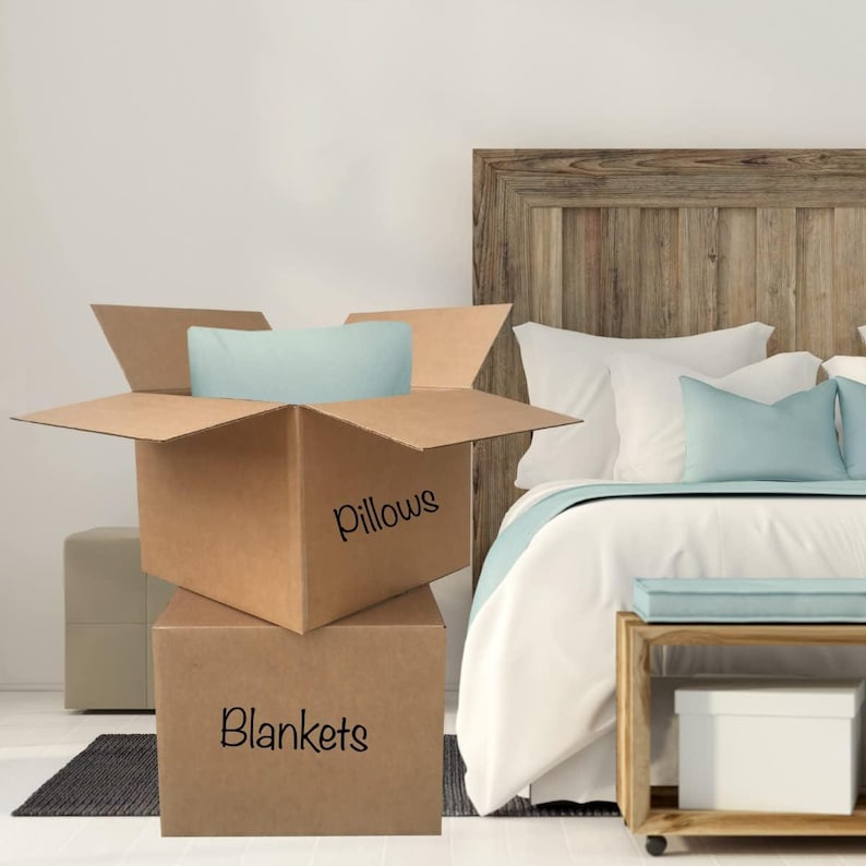 May include: Two cardboard boxes labeled "Pillows" and "Blankets" are stacked. The top box is open, revealing a light blue pillow. A bed with white bedding and light blue accents is in the background. A wooden headboard and nightstand are also visible.