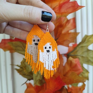 Puede incluir: Un par de pendientes de fantasma de cuentas naranja y blancas con ojos negros y un gancho plateado. Los pendientes cuelgan de una mano sobre un fondo de hojas de otoño naranja y marrón.