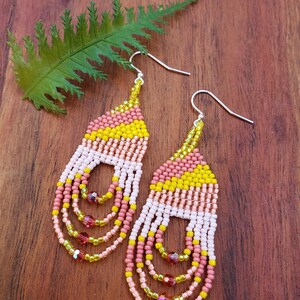 May include: A pair of long, beaded earrings with a pink, white, and yellow color scheme. The earrings have a fringe-like design with small crystal accents.