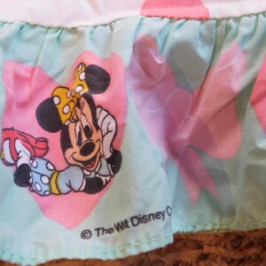 Puede incluir: Una tela azul claro y rosa con un ribete de volantes. La tela presenta una imagen de dibujos animados de Minnie Mouse con un lazo rojo y un coraz&oacute;n rosa. El texto "The Walt Disney Company" est&aacute; impreso en negro en la parte inferior de la tela.