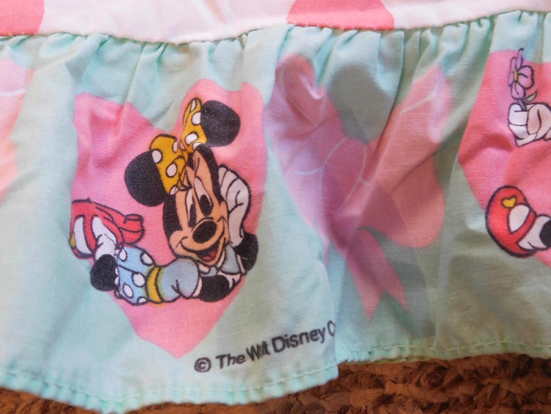 Puede incluir: Una tela azul claro y rosa con un ribete de volantes. La tela presenta una imagen de dibujos animados de Minnie Mouse con un lazo rojo y un coraz&oacute;n rosa. El texto "The Walt Disney Company" est&aacute; impreso en negro en la parte inferior de la tela.