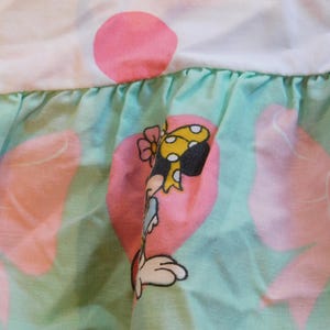 Puede incluir: Una tela verde y rosa con un ribete de volantes. La tela presenta un personaje de dibujos animados con un lazo amarillo y un coraz&oacute;n rosa.