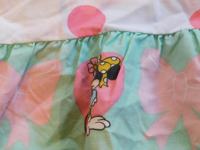 Puede incluir: Una tela verde y rosa con un ribete de volantes. La tela presenta un personaje de dibujos animados con un lazo amarillo y un coraz&oacute;n rosa.