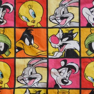 May include: A colorful fabric featuring a grid of cartoon character faces. The characters include Bugs Bunny, Daffy Duck, Tweety Bird, Sylvester the Cat, Pepe Le Pew, and Marvin the Martian, set against a background of pink, orange, and yellow squares.