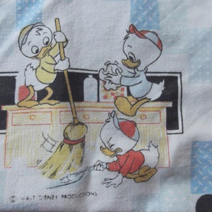 Pu&ograve; includere: Un tessuto bianco con un motivo ripetuto di tre paperi dei cartoni animati che puliscono una cucina. Un papero spazza con una scopa, un altro pulisce una superficie e il terzo pulisce il pavimento. Il testo Walt Disney Productions &egrave; stampato in basso.