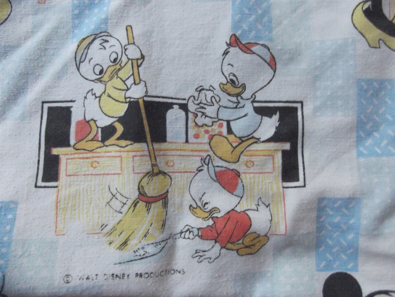Pu&ograve; includere: Un tessuto bianco con un motivo ripetuto di tre paperi dei cartoni animati che puliscono una cucina. Un papero spazza con una scopa, un altro pulisce una superficie e il terzo pulisce il pavimento. Il testo Walt Disney Productions &egrave; stampato in basso.