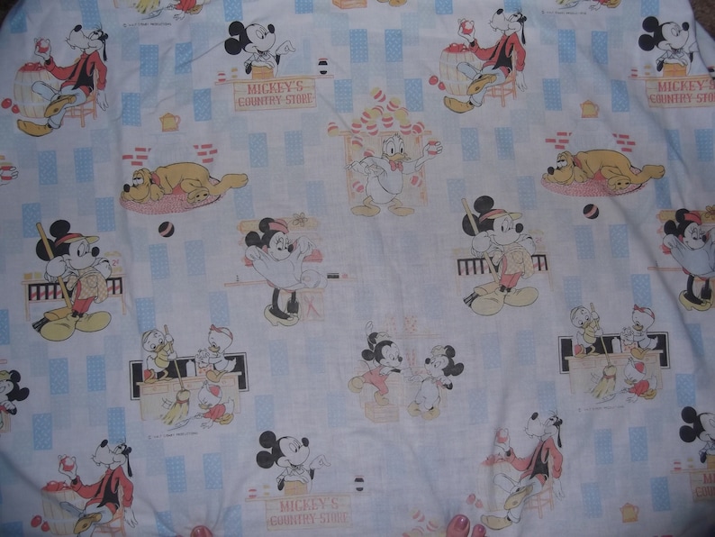 Pu&ograve; includere: Un tessuto bianco con un motivo ripetuto di personaggi Disney, tra cui Topolino, Minni, Paperino, Pippo e Pluto. La stampa include il testo Mickey's Country Store, con accenti rettangolari azzurri distribuiti su tutto il design.