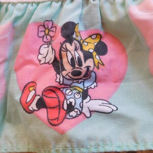 Puede incluir: Una tela azul claro con corazones rosas y una imagen de dibujos animados de Minnie Mouse sosteniendo una flor. Minnie Mouse lleva un vestido rojo con lunares blancos y un lazo amarillo.