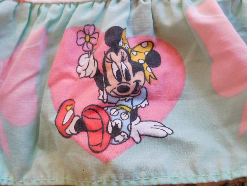 Puede incluir: Una tela azul claro con corazones rosas y una imagen de dibujos animados de Minnie Mouse sosteniendo una flor. Minnie Mouse lleva un vestido rojo con lunares blancos y un lazo amarillo.
